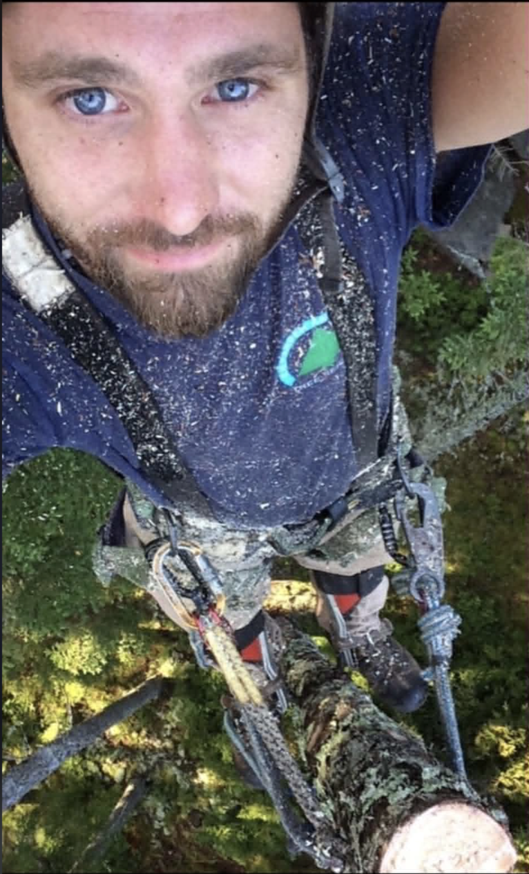 Owen Humphrey taking a selfie from high in a tree while wearing climbing gear and safety ropes